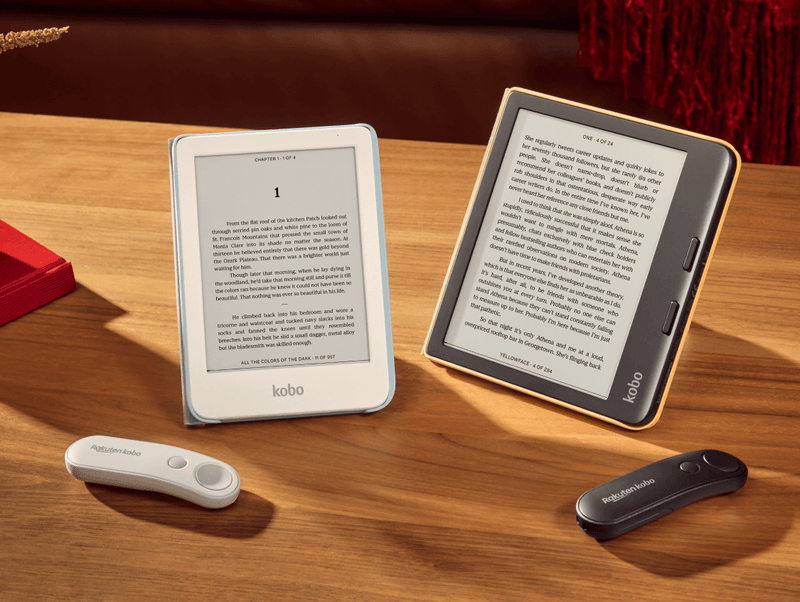 A white Kobo Clara Colour and black Kobo Libra Colour are propped up on a table in their SleepCovers with built-in stands, alongside two Kobo Remotes, one white and one black.