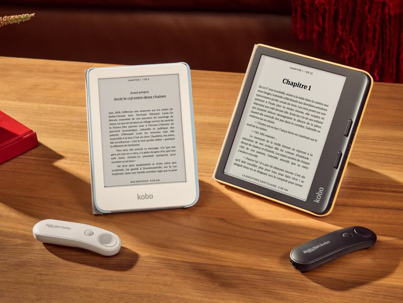 A white Kobo Clara Colour and black Kobo Libra Colour are propped up on a table in their SleepCovers with built-in stands, alongside two Kobo Remotes, one white and one black.
