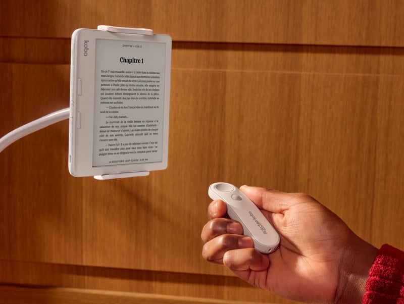 A white Kobo Remote is shown in someone's right hand, pointing towards a white Kobo Libra Colour eReader on a bendable stand.
