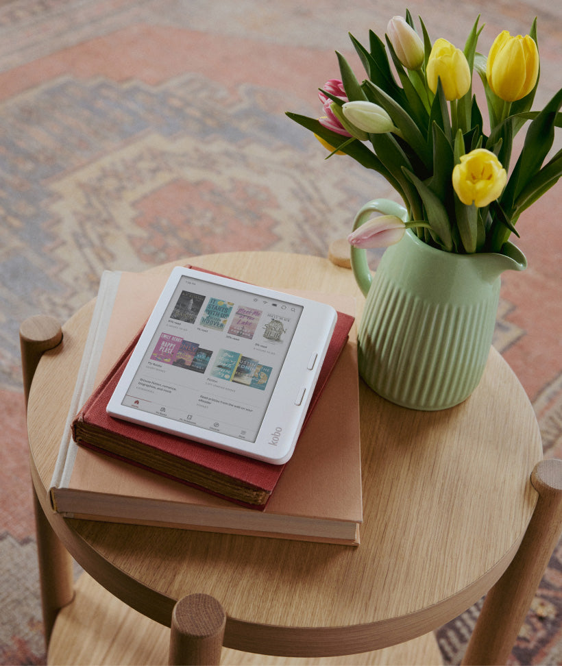 A Kobo Libra Colour eReader sitting on two hardcover books, set on a wooden table by flowers in a green vase.